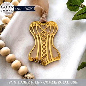 Könnte beinhalten: Ein goldfarbener Metallausschnitt eines Korsetts auf beigefarbenem Hintergrund. Der Ausschnitt ist mit filigranen Linien und Kurven verziert. Der Text "Salt & Smoke Laser Tested" ist in der oberen linken Ecke des Bildes sichtbar.