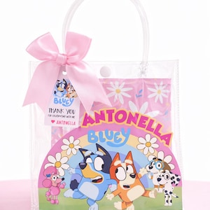 Puede incluir: Bolsa de regalo transparente con un lazo rosa y una etiqueta que dice "Thank You" con el nombre "Antonella". La bolsa presenta un gráfico colorido de los personajes de Bluey, un arcoíris y margaritas.