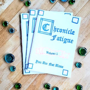 May include: Three white zines with blue and pink accents. The front cover of the top zine reads "Chronicle Fatigue" in blue cursive lettering with a blue "C" in a decorative frame. The text "Volume 1" is in pink and the text "You Are Not Alone" is in blue.