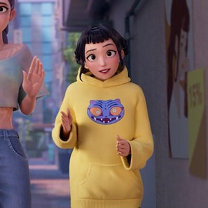 Puede incluir: Una persona con una sudadera con capucha amarilla con un gr&aacute;fico de gato de dibujos animados azul y morado. La sudadera tiene capucha y bolsillo delantero. La persona est&aacute; afuera.