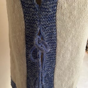 May include: Close-up of a cream-colored knitted vest with blue accents. The vest features a blue knitted trim and a blue tie closure at the neckline. The knitted fabric has a textured appearance.