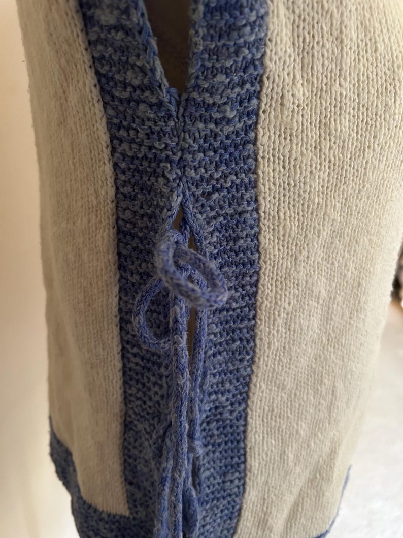 May include: Close-up of a cream-colored knitted vest with blue accents. The vest features a blue knitted trim and a blue tie closure at the neckline. The knitted fabric has a textured appearance.