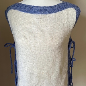 May include: A sleeveless, off-white knit top with a blue trim around the neckline and hem. Blue ties are on the sides. The top has a small embroidered detail on the front.