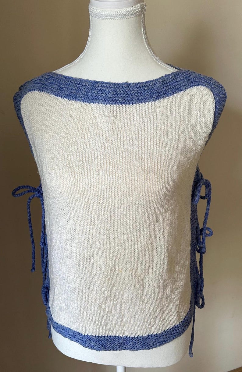 May include: A sleeveless, off-white knit top with a blue trim around the neckline and hem. Blue ties are on the sides. The top has a small embroidered detail on the front.