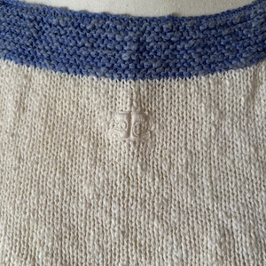 May include: Close-up of a cream-colored knit sweater with a blue knit neckline. The sweater has a textured appearance and a small embroidered emblem near the center. The neckline is a contrasting shade of blue.