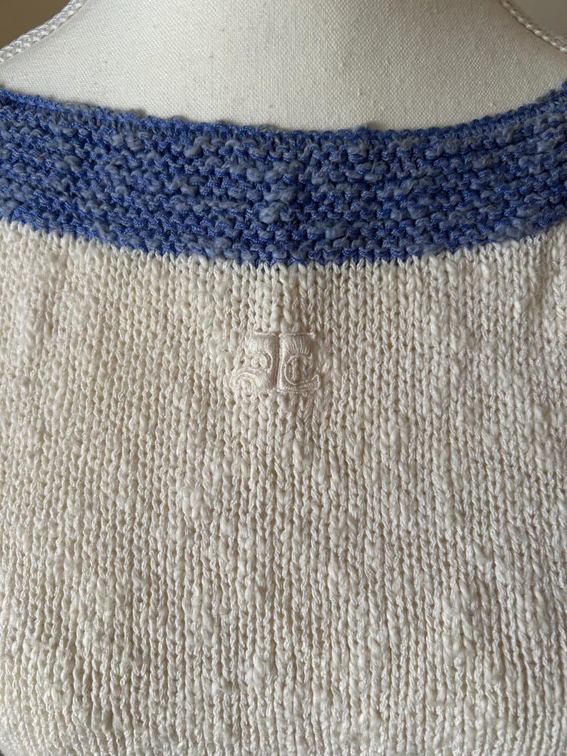 May include: Close-up of a cream-colored knit sweater with a blue knit neckline. The sweater has a textured appearance and a small embroidered emblem near the center. The neckline is a contrasting shade of blue.