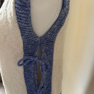 May include: A sleeveless, cream-colored knit top with blue trim and a laced front. The blue trim outlines the armholes and the front edges, with a blue tie closure. The knit fabric has a textured appearance.