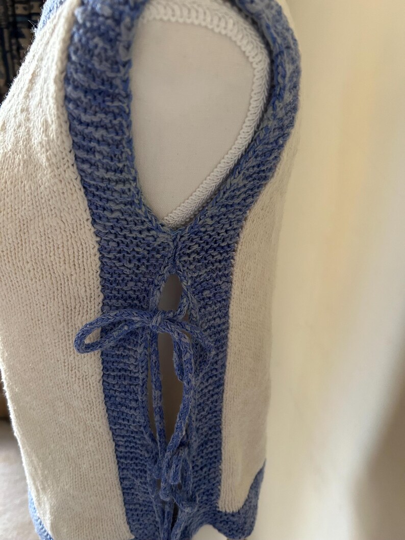 May include: A sleeveless, cream-colored knit top with blue trim and a laced front. The blue trim outlines the armholes and the front edges, with a blue tie closure. The knit fabric has a textured appearance.