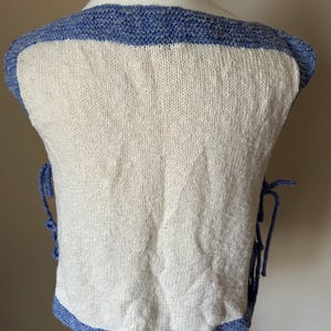 May include: A cream-colored knitted vest with a blue border and trim. The vest has blue ties on the sides and a square neckline. The knitted fabric has a textured appearance.
