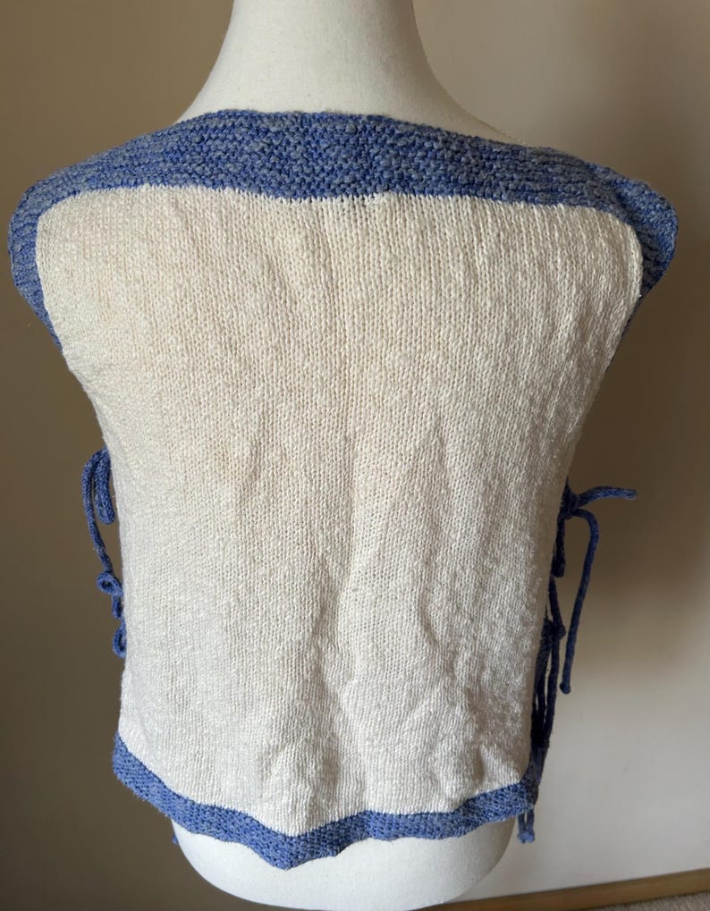 May include: A cream-colored knitted vest with a blue border and trim. The vest has blue ties on the sides and a square neckline. The knitted fabric has a textured appearance.