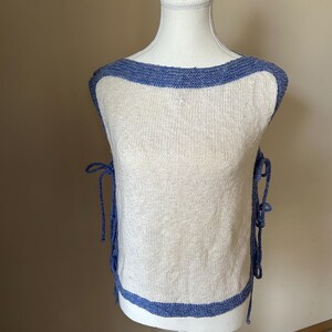 May include: A sleeveless, off-white knit top with a blue trim around the neckline and hem. The top has blue ties on each side. The garment is displayed on a white mannequin.