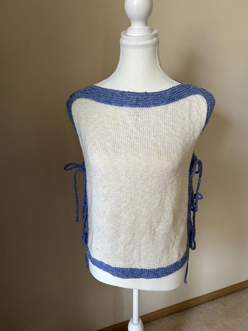 May include: A sleeveless, off-white knit top with a blue trim around the neckline and hem. The top has blue ties on each side. The garment is displayed on a white mannequin.