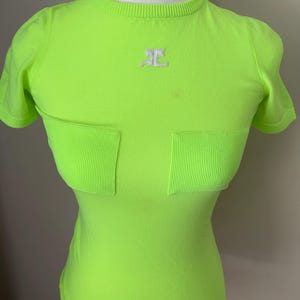 Puede incluir: Una camiseta de manga corta de color verde ne&oacute;n con un logotipo blanco en la parte delantera. La camiseta tiene dos bolsillos en la parte delantera.