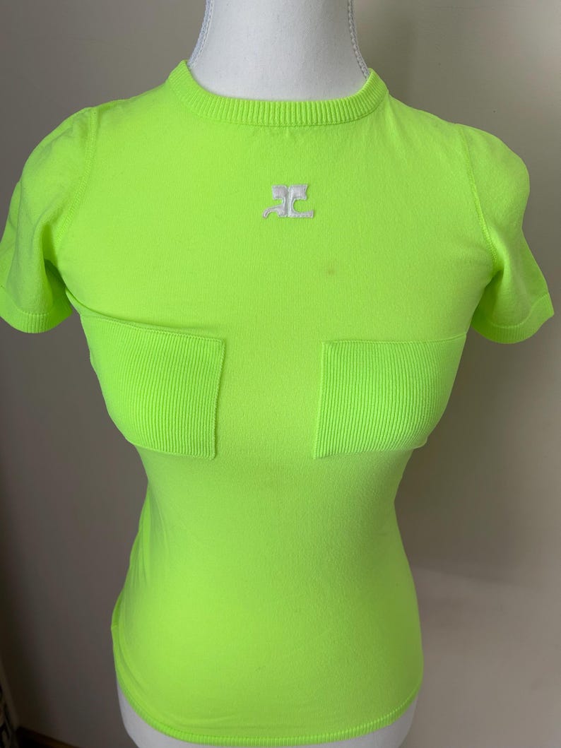 Puede incluir: Una camiseta de manga corta de color verde ne&oacute;n con un logotipo blanco en la parte delantera. La camiseta tiene dos bolsillos en la parte delantera.