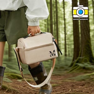 Peut inclure: Un sac d'appareil photo beige avec une bandoulière et une poignée. Le sac a une fermeture éclair noire et un logo noir qui dit "Marry IVI". Le sac est tenu dans un cadre forestier.