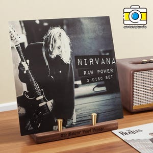 以下が含まれることがあります： 木製のスタンドに飾られた、タイトル「Raw Power 3 Disc Set」のニルヴァーナの白黒アルバムカバー。アルバムにはギターを演奏する人の写真が掲載されています。小さな茶色のスピーカーとビートルズのアルバムも表示されています。