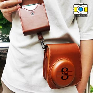 以下が含まれることがあります： 茶色の革製カメラケースと、お揃いのカードホルダー。 カメラケースの前面は丸く、「Mui」という名前がプリントされています。 カードホルダーはストラップと金属製の留め金でカメラケースに取り付けられています。 「camera accessories」の文字が右上に印刷されています。