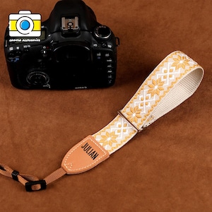 Könnte beinhalten: Eine Kamerahandschlaufe mit einem weißen und goldenen Blumenmuster. Der Riemen hat ein Lederende mit dem Aufdruck "JULIAN". Im Hintergrund befindet sich eine schwarze Kamera.