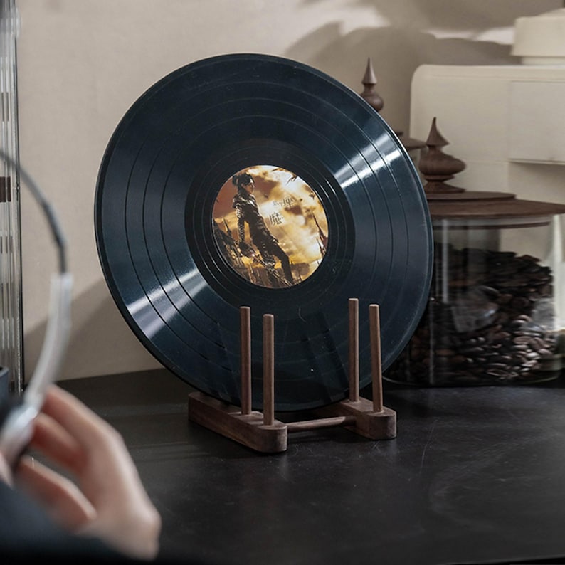 May include: A black vinyl record displayed on a wooden stand. The record has a colorful label with an image of a person. The stand is made of dark wood and has a simple, minimalist design. The record is displayed on a dark surface.