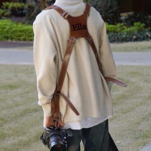 以下が含まれることがあります： 背面には「Ella」の文字がエンボス加工された、茶色の革製カメラハーネス。ハーネスは調節可能なストラップと金属製の金具が付いています。カメラマンは黒いカメラを持っています。