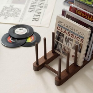 May include: A wooden CD and vinyl record holder. The holder is dark brown and holds several CDs and vinyl records. The CDs are in clear cases, and the vinyl records are black with colorful labels. The text "BROOKS & DUNN CLASSICS" is visible.