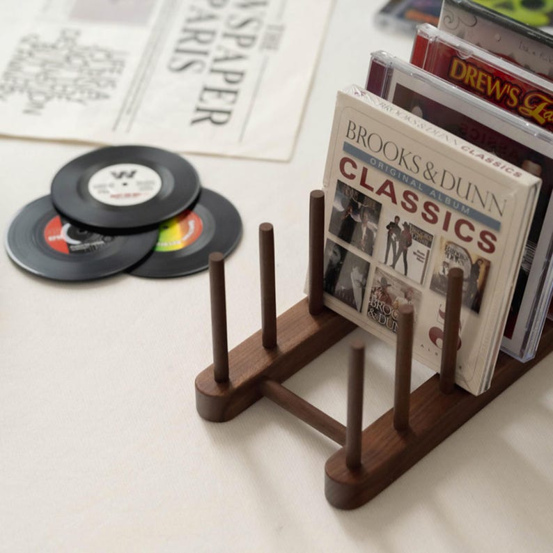 May include: A wooden CD and vinyl record holder. The holder is dark brown and holds several CDs and vinyl records. The CDs are in clear cases, and the vinyl records are black with colorful labels. The text "BROOKS & DUNN CLASSICS" is visible.