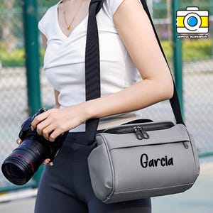 May include: A gray camera bag with a black shoulder strap. The bag has a zippered top and a front pocket. The word "Garcia" is printed on the front of the bag. The image also shows a camera and the text "camera accessories".