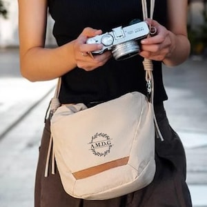 May include: A cream-colored camera bag with a brown accent and a shoulder strap. The bag has a circular emblem with the text "A.M.D.G." inside a wreath. A silver camera is held in the background.