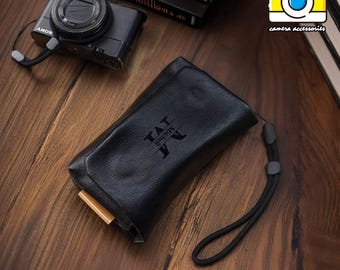 May include: A black leather camera case with a personalized inscription that reads "TAT BROWN JA". The case has a black strap and a wooden zipper pull.