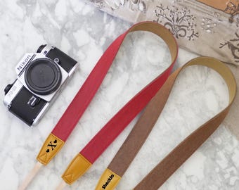 May include: A silver and black Nikon camera with three leather camera straps in red, brown, and tan. The straps have tan leather ends with the text "X" and "Shecklo" printed on them. The straps are on a marble surface.