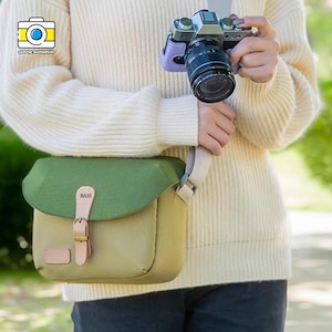 Könnte beinhalten: Eine grüne und beige Kameratasche mit Klappe und hellbraunem Lederriemen. Die Tasche hat einen Schnallenverschluss und die Initialen "R&H" sind sichtbar. Eine Kamera wird im Hintergrund gehalten.