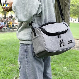 May include: A grey camera bag with a black strap and trim is worn over the shoulder. The bag features a front pocket and a top flap. The name "Ekkert" is printed on the front, along with the brand name "boona".