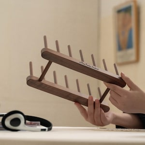 May include: A wooden thread rack with multiple pegs, held in front of a neutral background. The rack is made of dark wood and has a minimalist design. The text "by Wilson Family & Soundtrack" is printed on the rack.