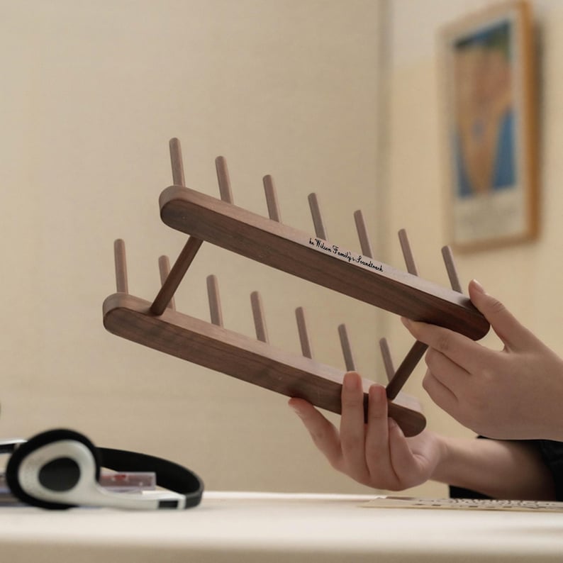 May include: A wooden thread rack with multiple pegs, held in front of a neutral background. The rack is made of dark wood and has a minimalist design. The text "by Wilson Family & Soundtrack" is printed on the rack.