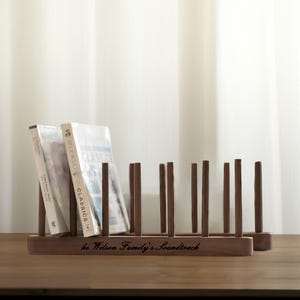 May include: A dark brown wooden CD or DVD rack with vertical dividers. The rack holds several CDs or DVDs, with the text "he Wilson Family's Soundtrack" engraved on the front. The rack is on a wooden surface.