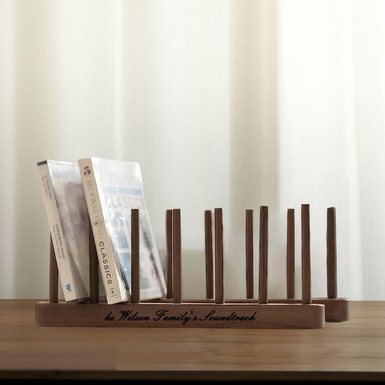 May include: A dark brown wooden CD or DVD rack with vertical dividers. The rack holds several CDs or DVDs, with the text "he Wilson Family's Soundtrack" engraved on the front. The rack is on a wooden surface.