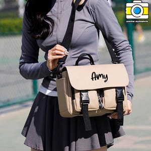 Könnte beinhalten: Beige Kameratasche mit schwarzen Riemen und Schnallen. Die Tasche hat zwei Vordertaschen und einen oberen Griff. Der Name "Amy" steht in schwarzer Schrift auf der Vorderseite. Für Kamerazubehör.