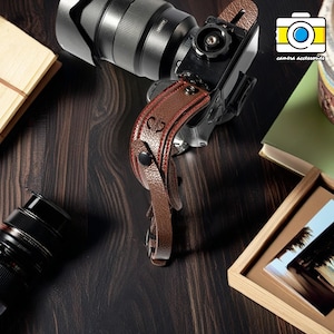 May include: A brown leather camera strap with a red stitching detail and a metal button. The strap is attached to a black camera with a lens.