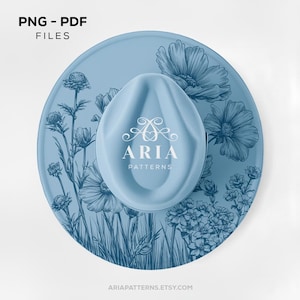 May include: A light blue hat with a floral design and the text "ARIA PATTERNS". The hat is round with a curved top and features a detailed botanical illustration in a darker shade of blue. The words "PNG - PDF FILES" are in the top left corner.