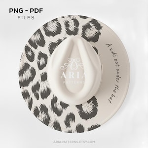 May include: A fedora hat with a cream-colored crown and a wide brim featuring a black and white leopard print. The brim has the text "A wild cat under the hat" and the Aria Patterns logo.