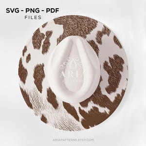 May include: A white cowboy hat with a brown and white cowhide pattern. The hat has a white crown and brim. The text "SVG - PNG - PDF FILES" is at the top, and "ARIAPATTERNS.ETSY.COM" is at the bottom.
