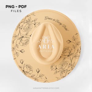 May include: A beige fedora hat with a cream-colored crown. The hat features a floral rose design in black ink around the brim. The text "Grace in Every Petal" and "ARIA PATTERNS" are printed on the hat.
