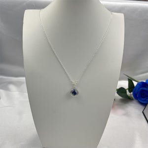 Può includere: Una collana d'argento con un piccolo pendente rotondo con una pietra blu scuro. Il pendente è circondato da un alone di pietre più piccole e trasparenti. La collana è esposta su un manichino bianco.