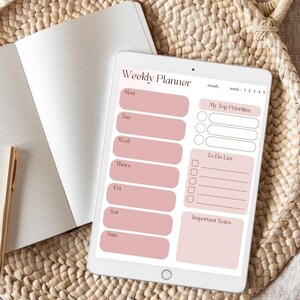 Op de afbeelding: Een digitale weekplanner op een tablet met een roze en wit ontwerp. De planner heeft secties voor elke dag, een takenlijst en ruimte voor notities. Een notitieboek en pen staan op de achtergrond. De tekst "Weekly Planner" is zichtbaar.