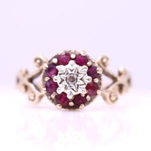 Könnte beinhalten: Ein Goldring mit einer zentralen Anordnung tiefroter Rubine in kreisförmiger Form. In der Mitte der Rubine befindet sich ein silberfarbenes, sternförmiges Element mit einem kleinen Stein. Die Ringschiene hat beidseitig dekorative Verzierungen.