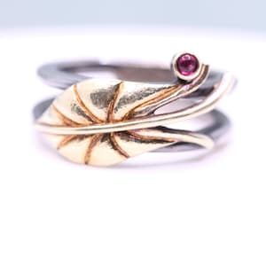 Könnte beinhalten: Ein silberner Ring mit einem goldenen Blattdesign und einem kleinen, runden, roten Edelstein. Das Blatt hat strukturierte Linien, und der Ring hat ein Doppelband. Der Edelstein ist auf einem zarten Zweig gesetzt.