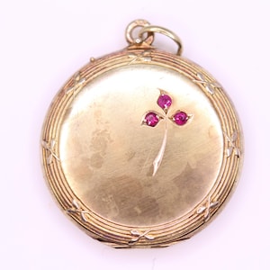 Può includere: Un ciondolo rotondo color oro con un motivo floreale. Il ciondolo presenta un design a trifoglio con tre gemme rosse. Il ciondolo ha un bordo strutturato e scanalato e un piccolo anello nella parte superiore per una catena.