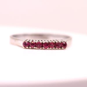 Può includere: Un anello d'argento con sei piccole pietre preziose rotonde di colore rosso intenso. Le pietre sono incastonate in una fila sulla parte superiore dell'anello. La fascia è di design semplice e piatto. L'anello è mostrato su sfondo bianco.