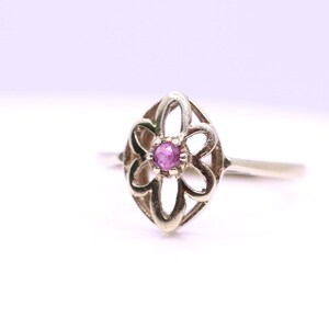 Può includere: Un anello in oro con un design floreale traforato a forma di marquise. Il centro del fiore è incastonato con una piccola pietra preziosa rosa, rotonda e sfaccettata, tenuta in posizione da quattro griffe. Il gambo è sottile e liscio.