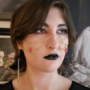 May include: Gold-toned face jewelry with a chain design. The piece drapes across the nose and features dangling red teardrop-shaped beads. Long chain earrings with a spike at the end complete the look. The jewelry is worn over a black top.